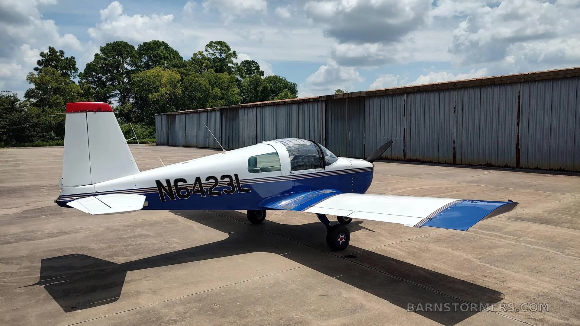 A white and blue American Aviation AA-1 Yankee aircraft with registration N6423L, viewed from the rear-left, parked on an asphalt surface next to a hangar.