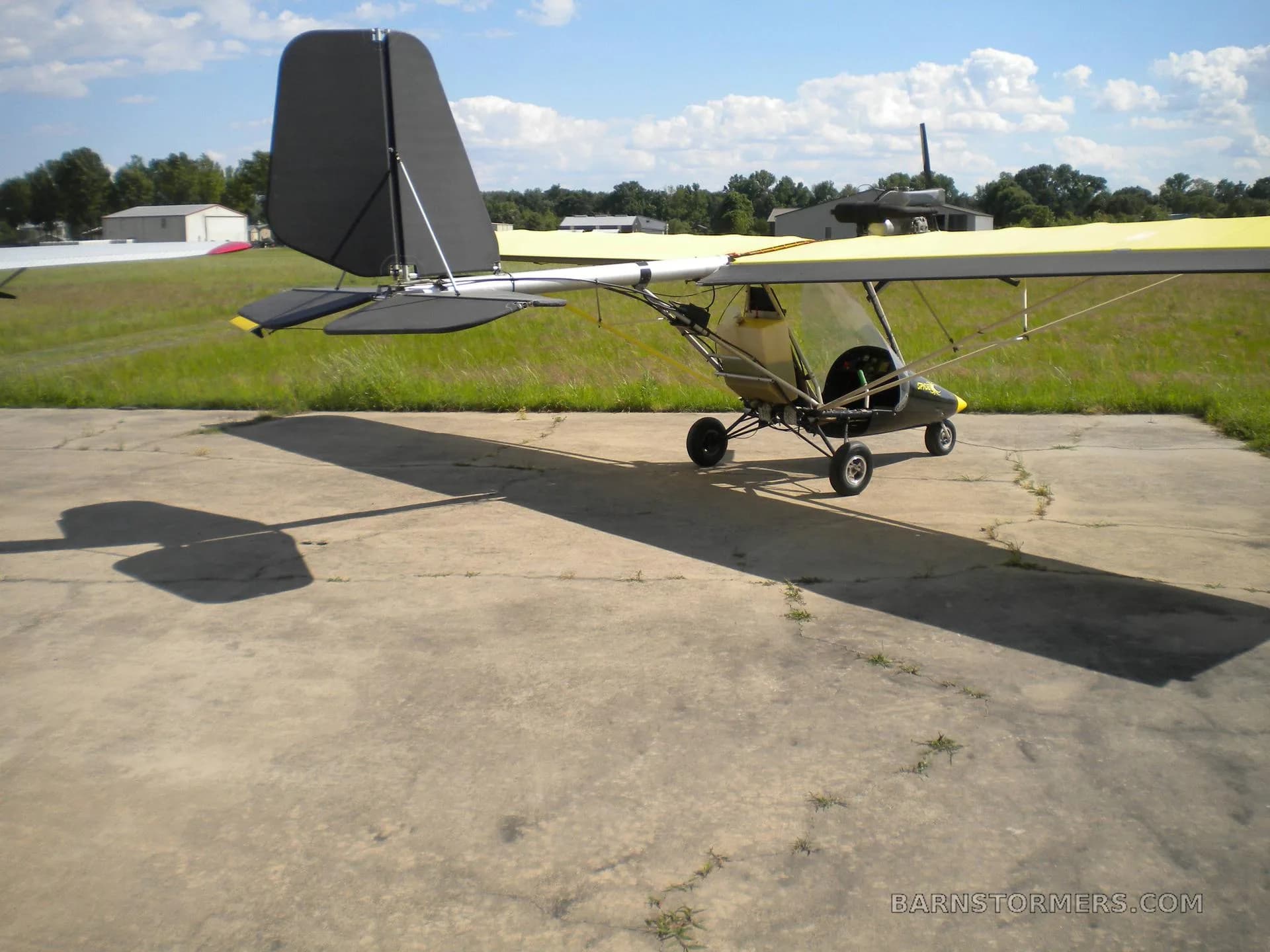 A yellow-winged ultralight aircraft with a black tail and nose cone, parked on a concrete pad at an airfield.
