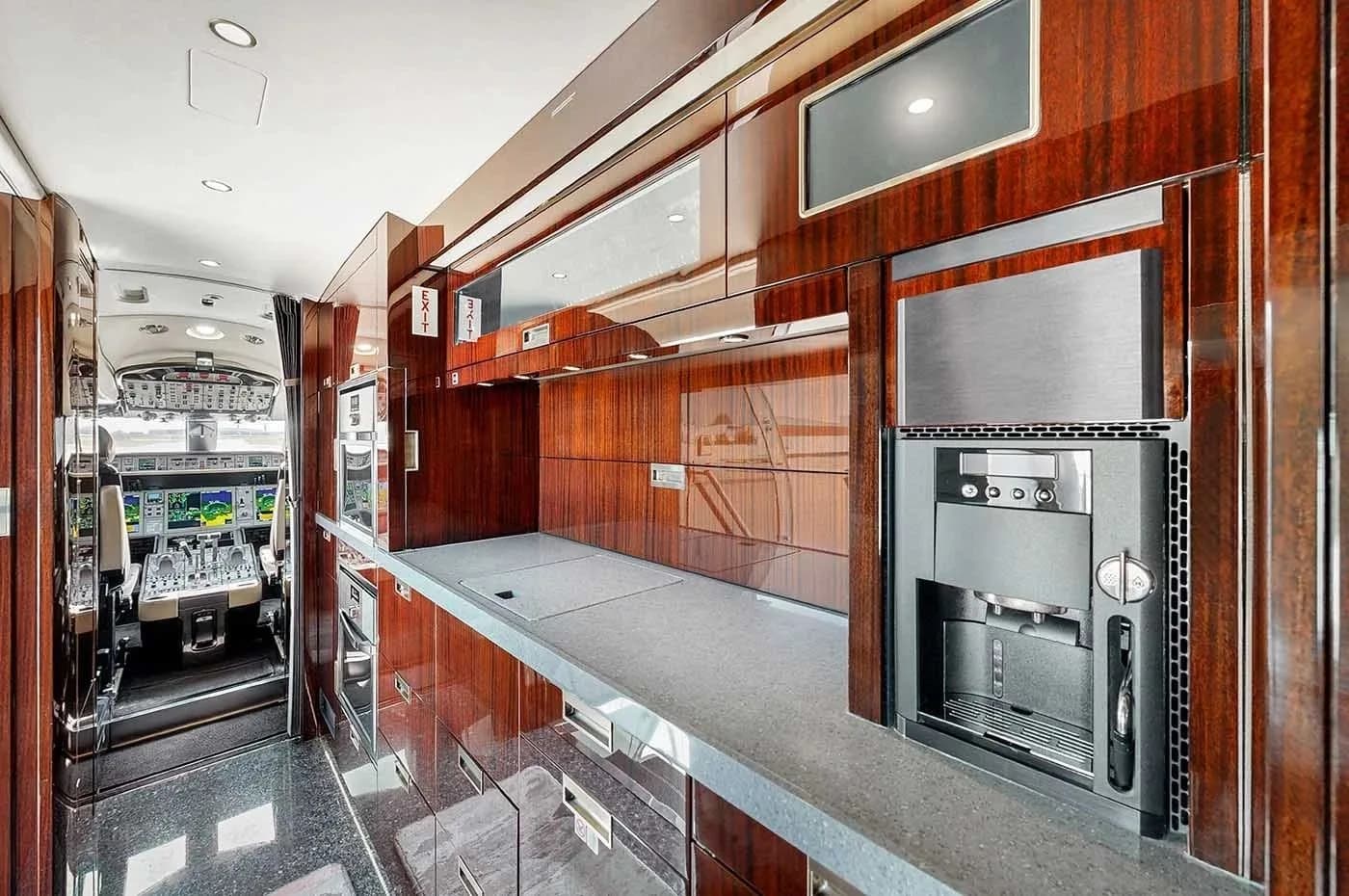 The galley of a private jet, featuring extensive dark wood cabinetry, a long countertop, and a built-in coffee machine, with the cockpit visible in the background.