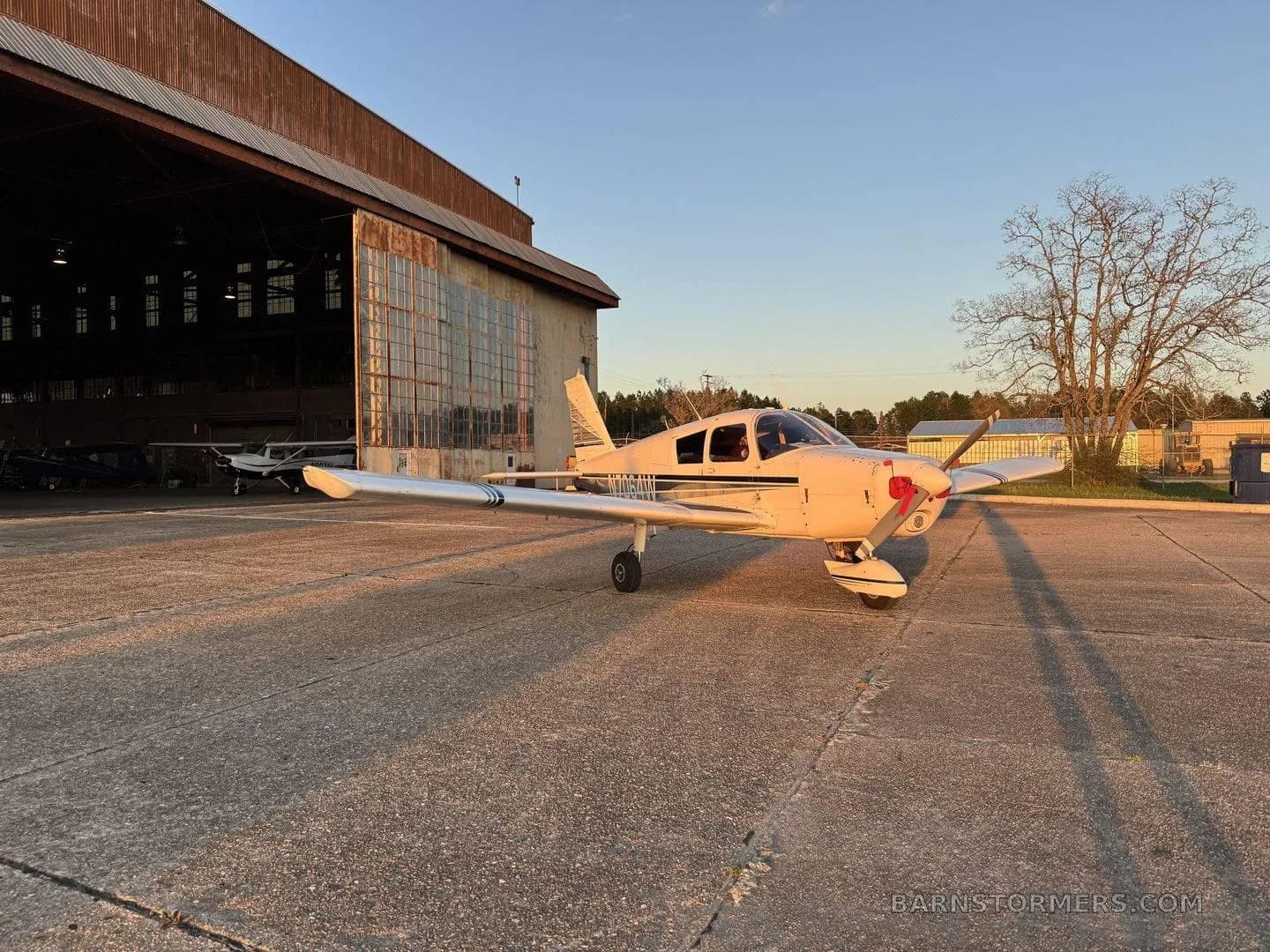 A white single-engine propeller aircraft with dark stripes parked on a concrete ramp with a hangar and trees in the background at sunset.