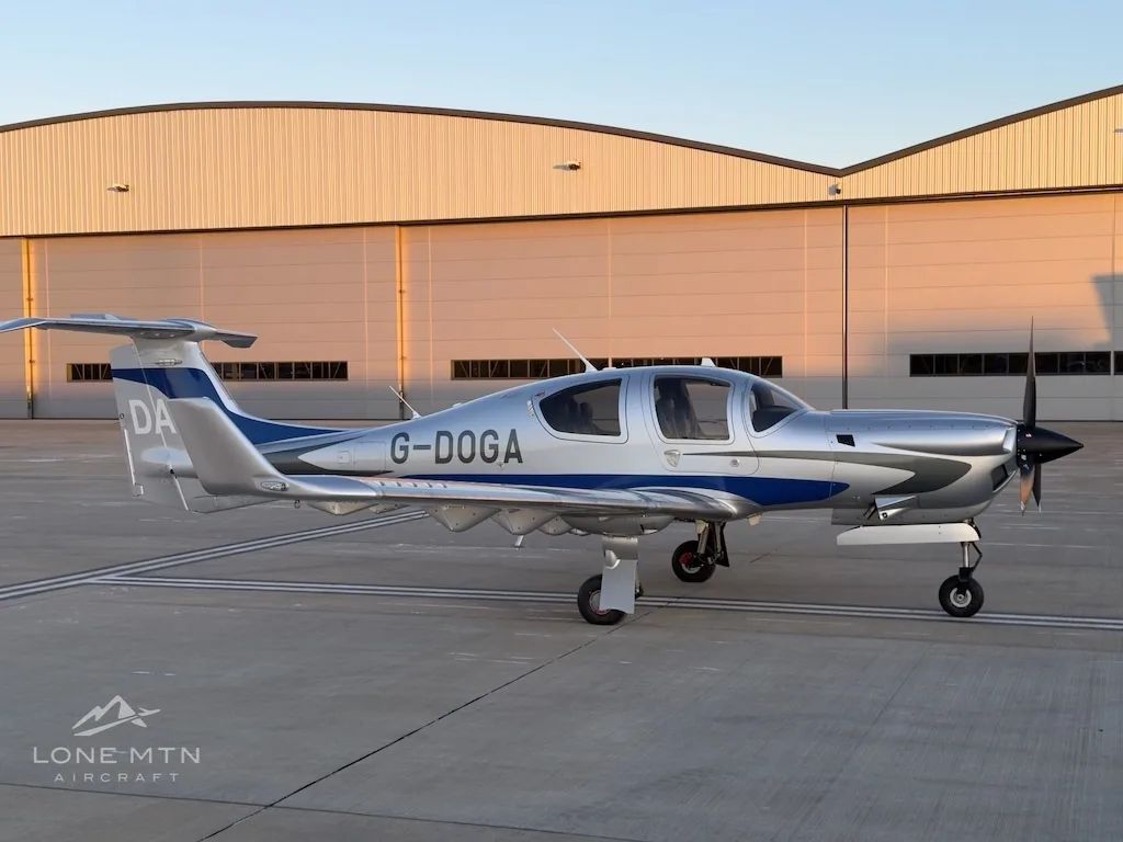 A slightly angled side view of a silver and blue Diamond DA50 aircraft with registration G-DOGA on an airfield.