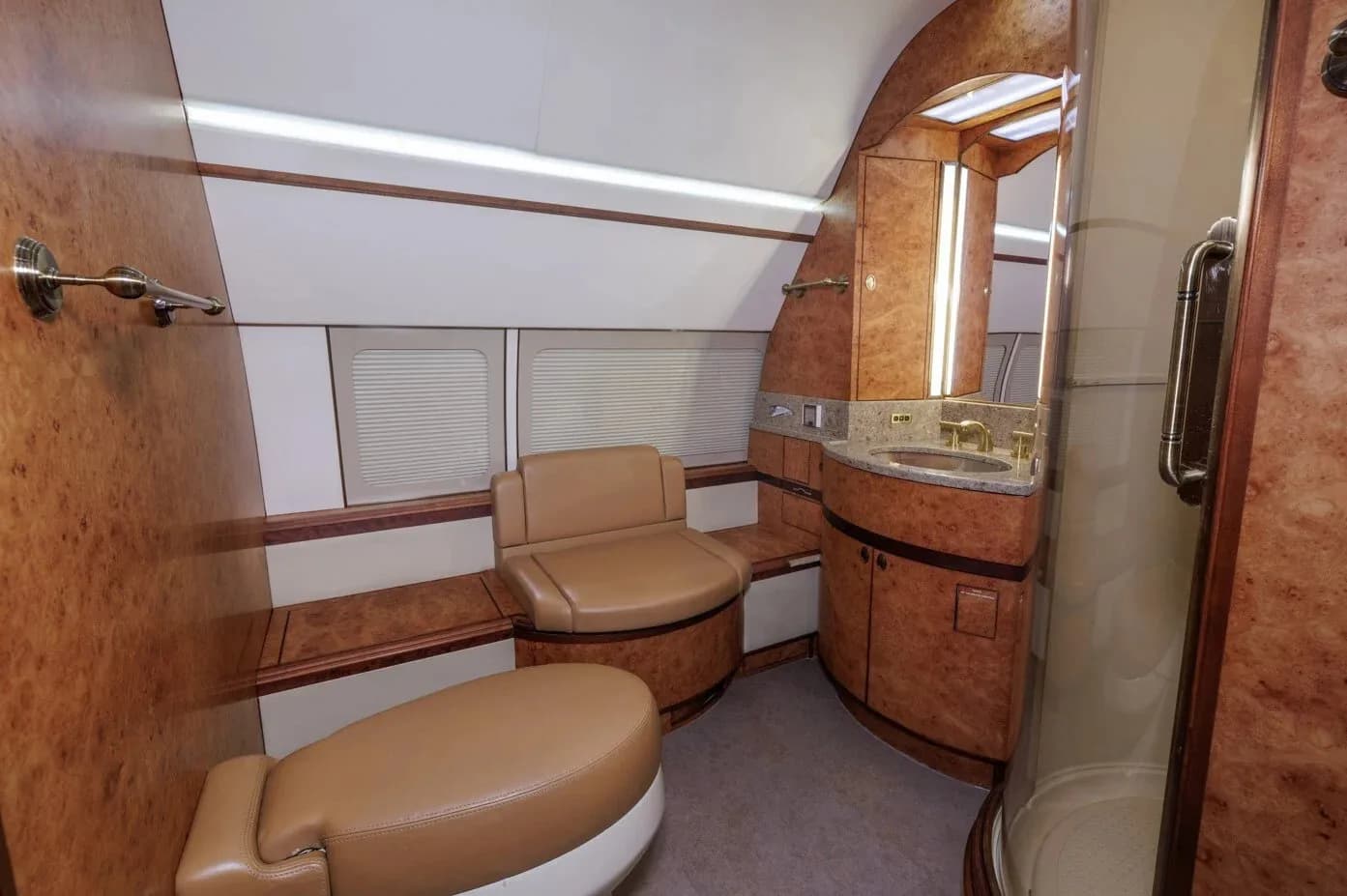 A luxurious aircraft lavatory with a sink, mirror, toilet, and shower, all finished with wood veneer.