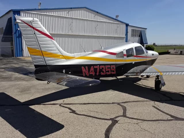 A silver, black, red, and yellow single-engine propeller aircraft, N47359, parked on an asphalt ramp next to a hangar.