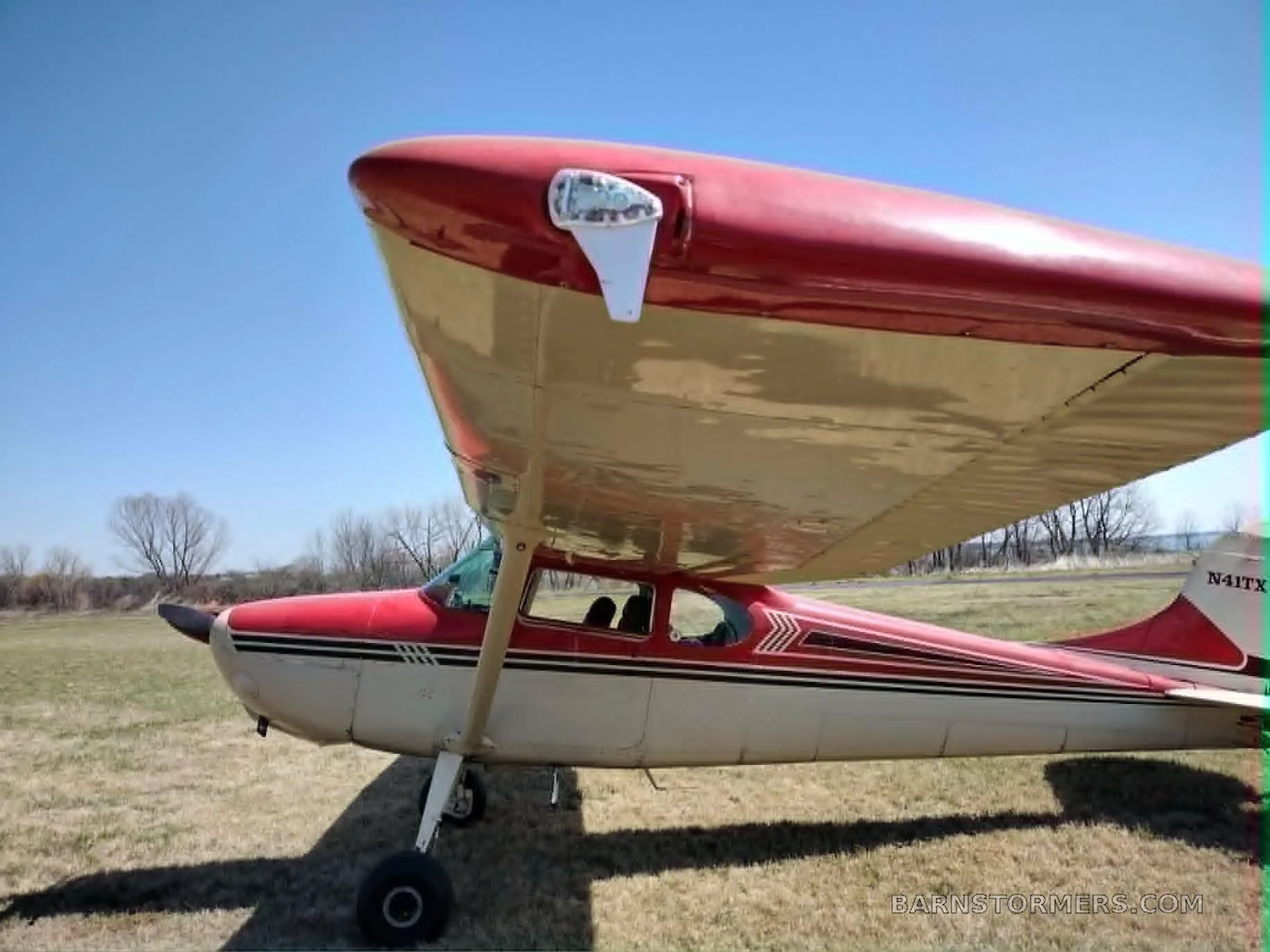 A side view of a red and white high-wing single-engine aircraft with registration N41TX, parked on a dry grassy field.