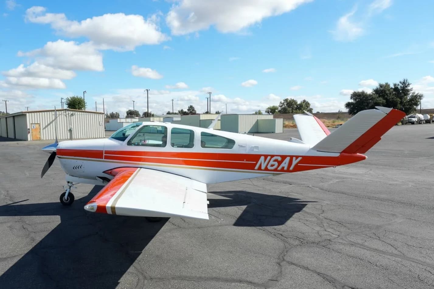 A white and orange Beechcraft Bonanza N6AY parked on an airport tarmac under a partly cloudy sky.