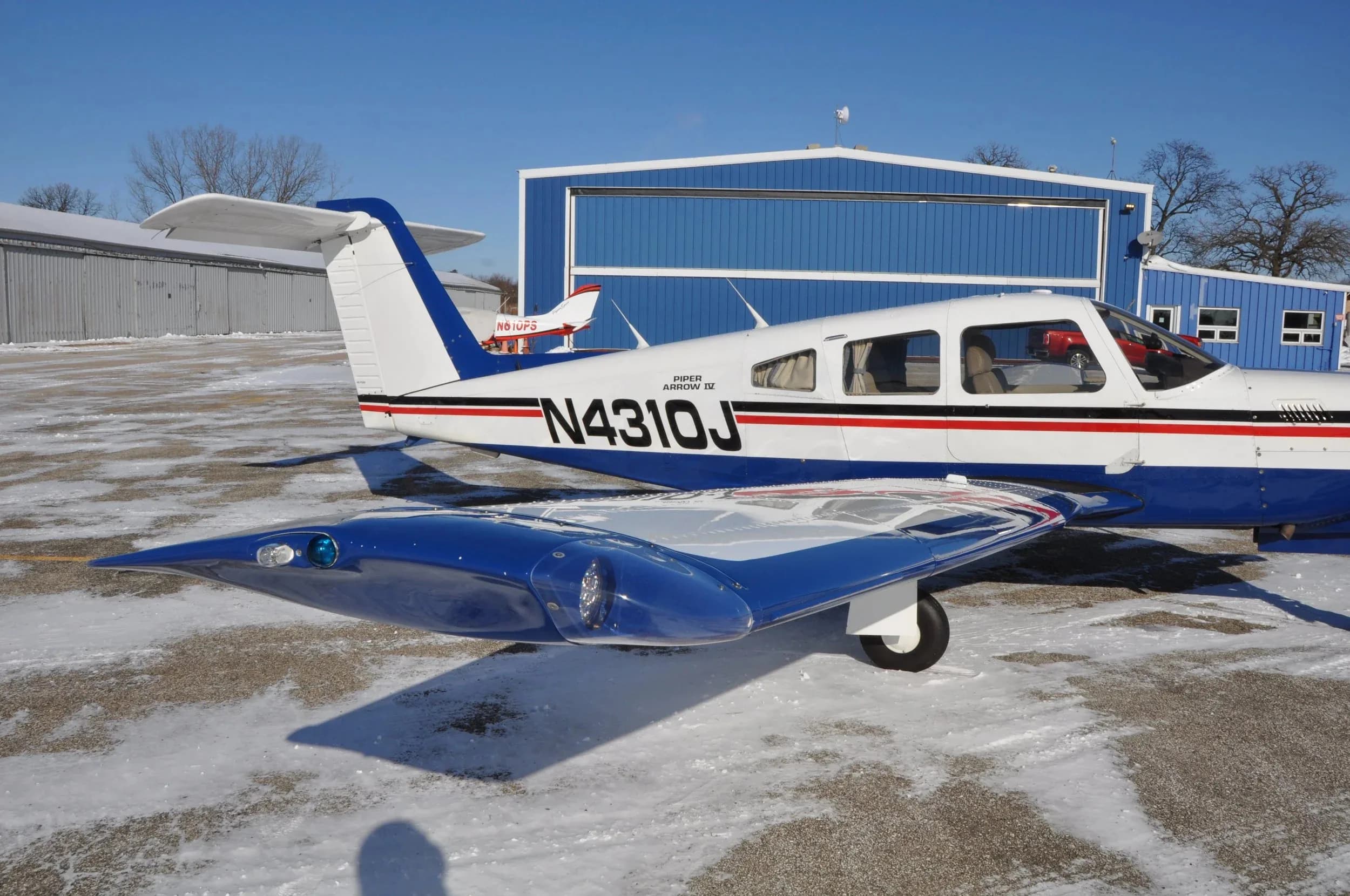 A close-up view of the right wing and fuselage of a white and blue Piper Arrow IV aircraft with registration N4310J on a snowy tarmac.