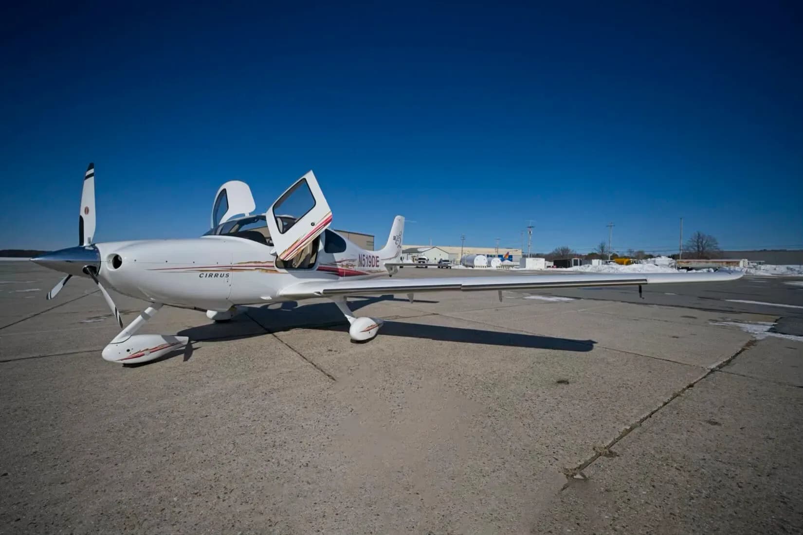 A wide-angle view of a white Cirrus SR22 aircraft with red and gold stripes, registration N519DE, parked on an airfield with its doors open.