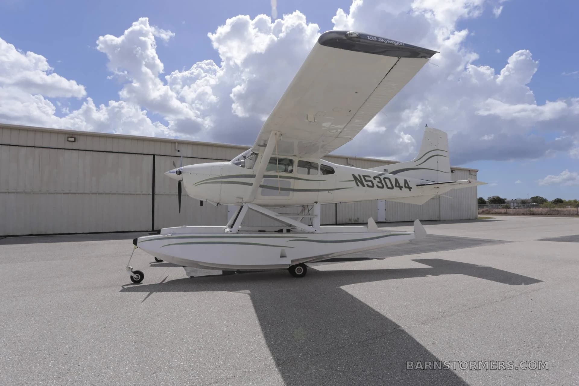 A white Cessna 180 Skywagon seaplane with green stripes and registration N53044 is parked on an airfield under a cloudy sky.