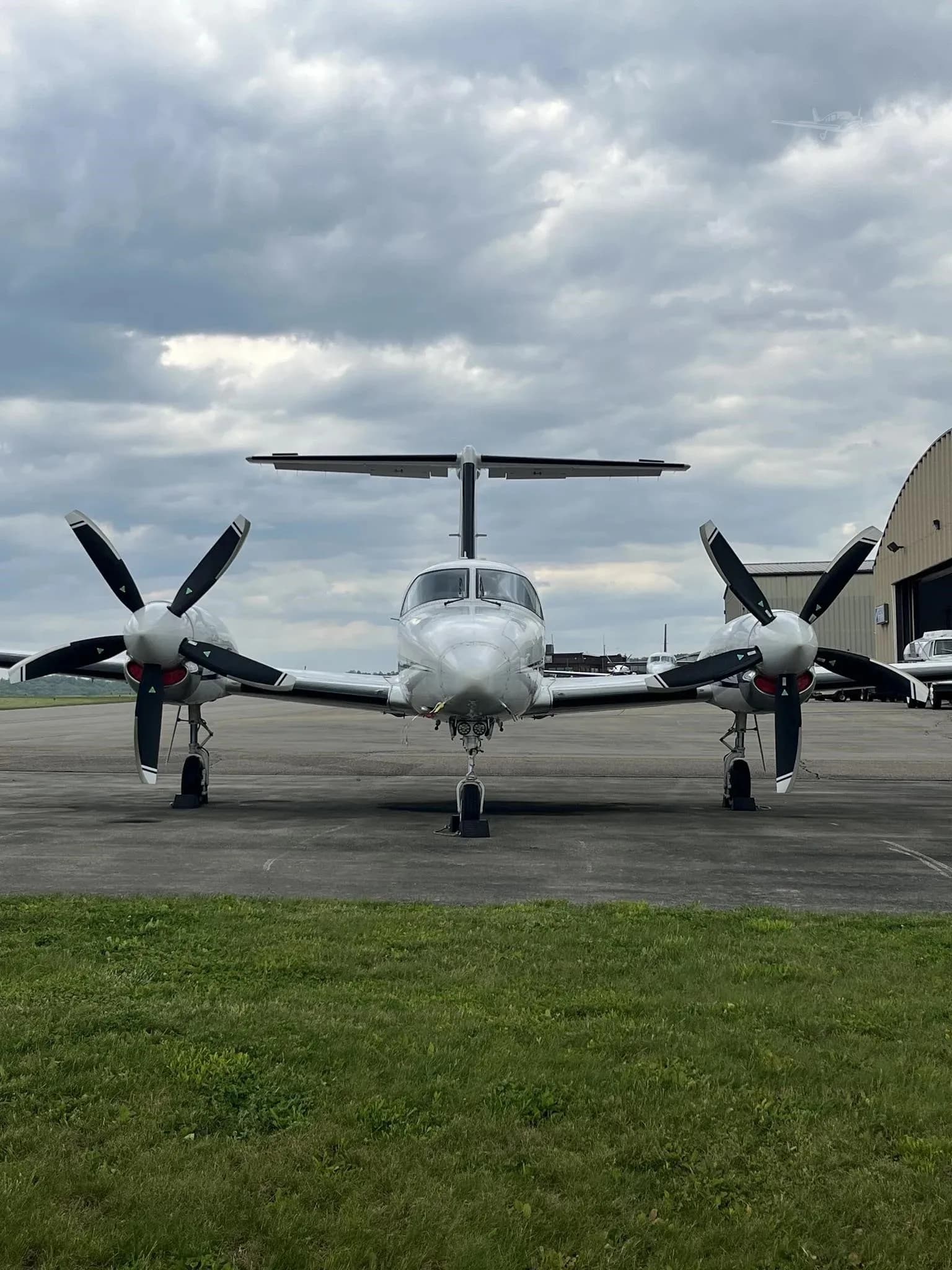Front view of a white twin-engine turboprop aircraft with black propellers on an airfield under a cloudy sky.