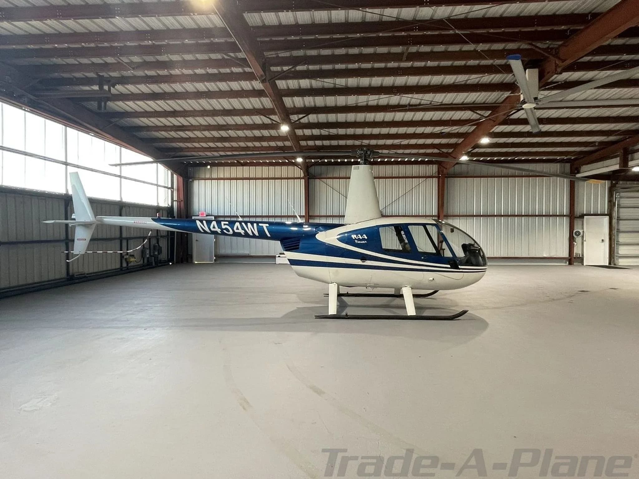 A blue and white Robinson R44 Raven helicopter with registration N454WT is parked inside a hangar, viewed from the front-left.