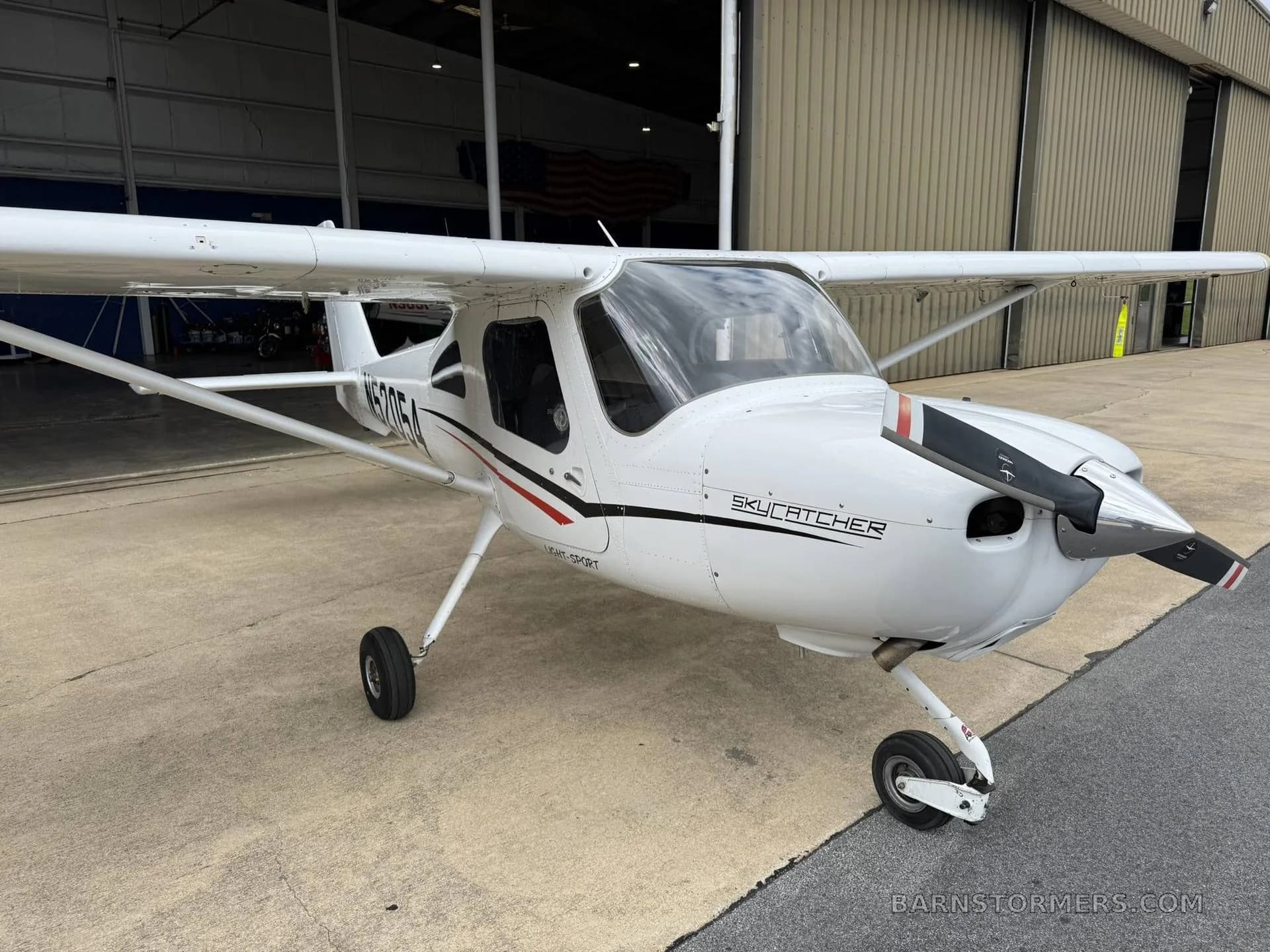 A white Cessna Skycatcher light-sport aircraft with black and red stripes, viewed from the front-right, parked outside a hangar.
