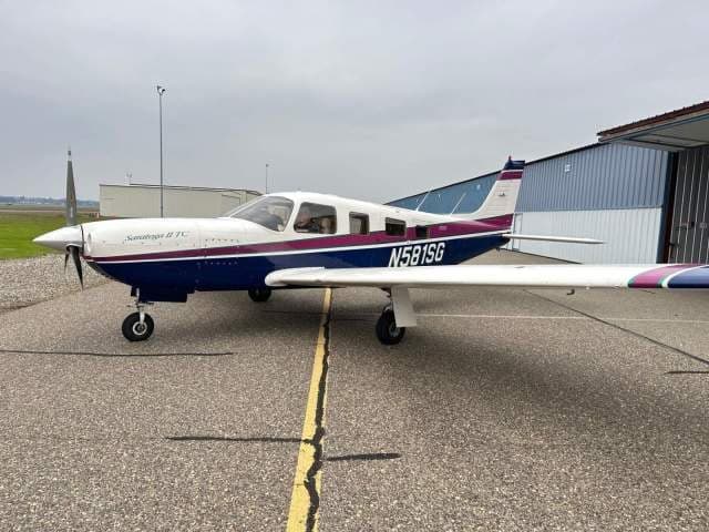 A blue, white, and purple Piper Saratoga II TC aircraft (N581SG) parked on an airfield tarmac.