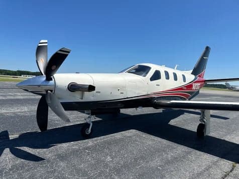 A front-side view of a white, red, and black single-engine turboprop aircraft with its propeller visible, parked on an airfield.