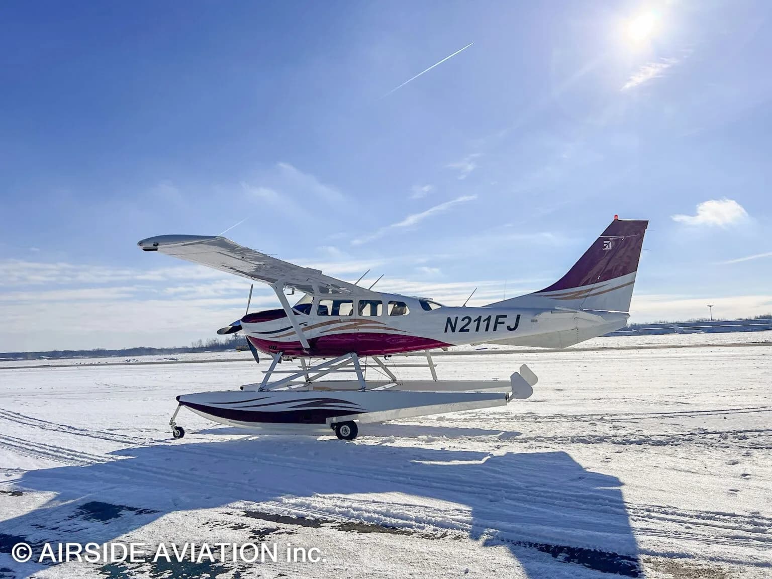 A red and white floatplane, N211FJ, is parked on a snow-covered field under a bright blue sky.