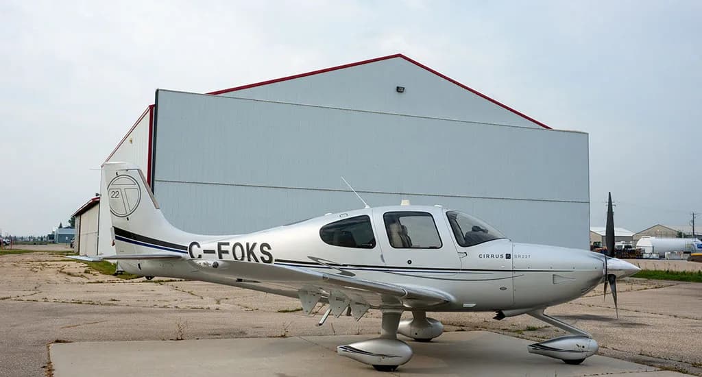 A silver Cirrus SR22T aircraft with registration C-FOKS is parked on an airfield in front of a white hangar.