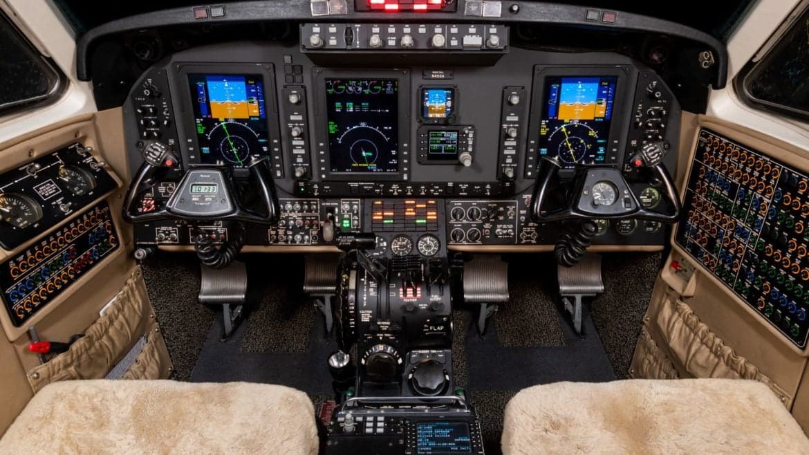 A detailed view of a modern aircraft cockpit featuring multiple digital displays, control yokes, and a central throttle quadrant.