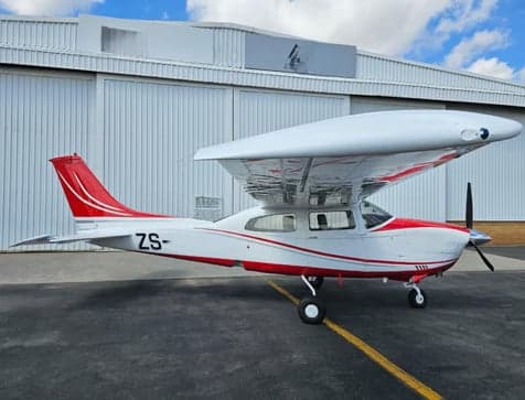 A white and red Cessna 210 Centurion aircraft with ZS- registration is parked on an airfield in front of a white hangar.