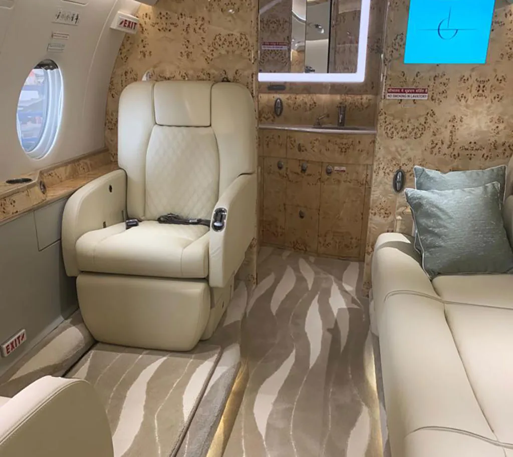 A close-up of a cream leather seat and a side sofa in a private jet cabin, with a small lavatory or galley area visible.