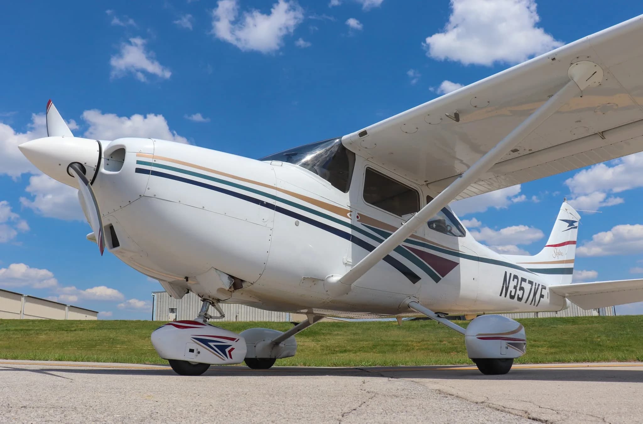 A low-angle front-quarter view of a white Cessna 182 Skylane with multi-color stripes on an airport ramp.