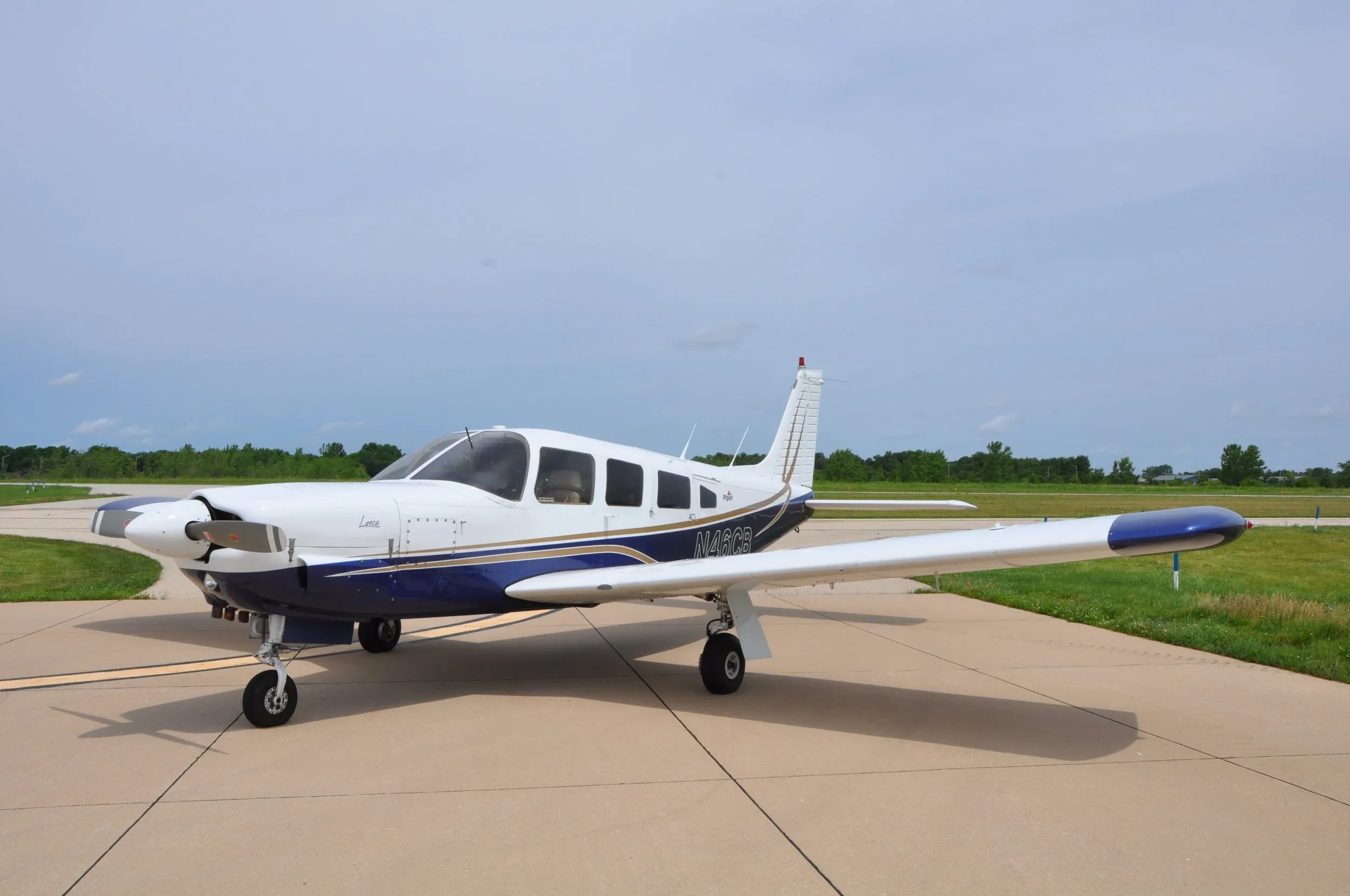 A white and blue Piper Lance aircraft (N46CB) parked on an airfield under a cloudy sky.