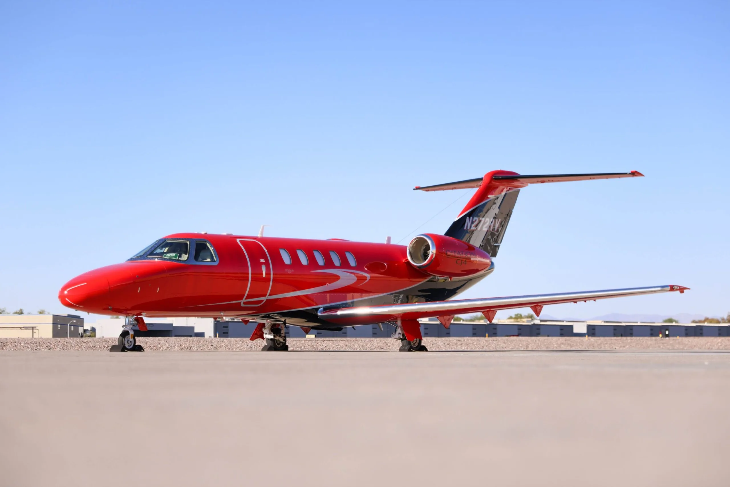 A red and black business jet, likely a Citation CJ4, is parked on an airfield under a clear blue sky.