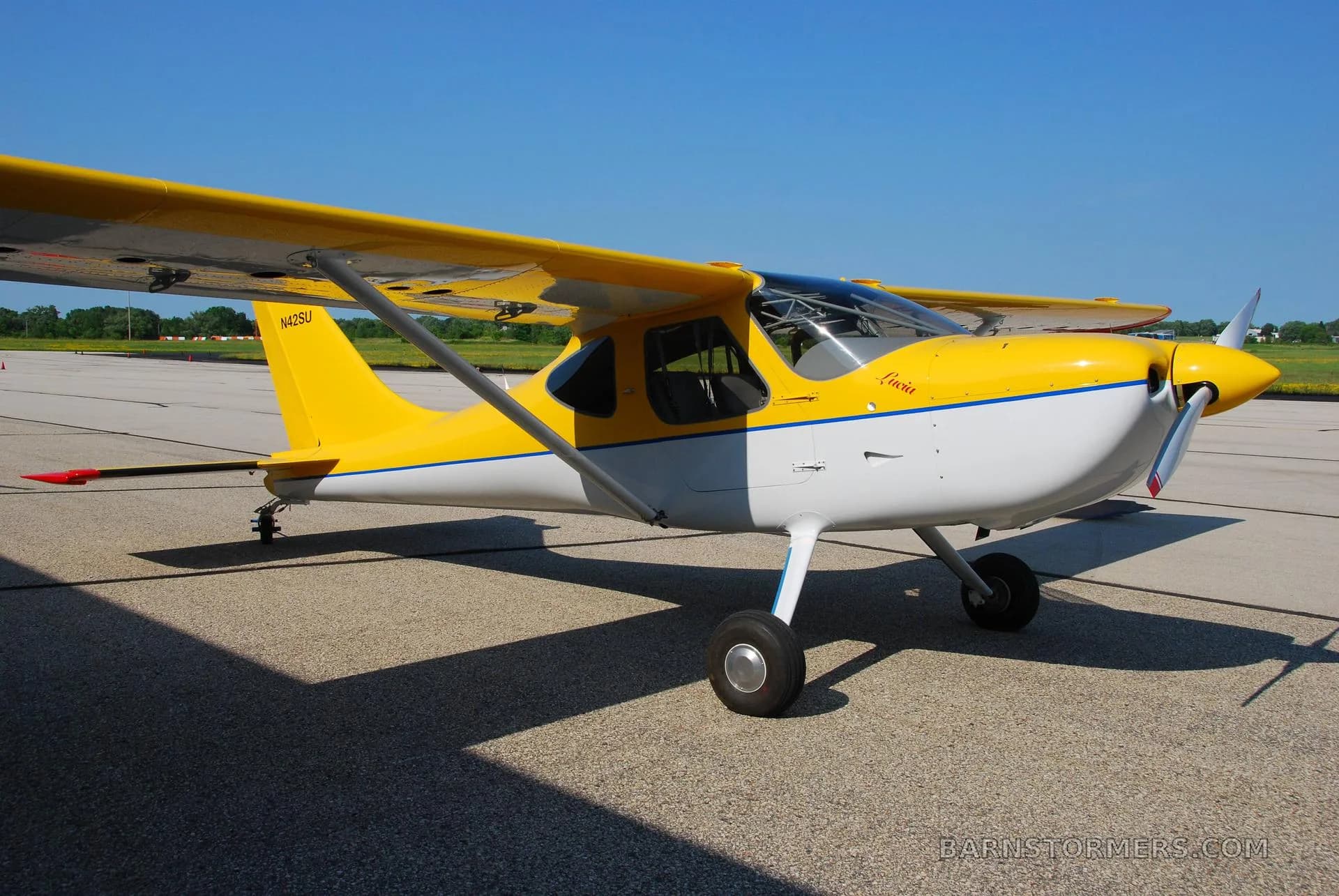 A yellow and white light sport aircraft with a blue stripe, registration N42SU, parked on an asphalt runway under a clear blue sky, viewed from the front-right.