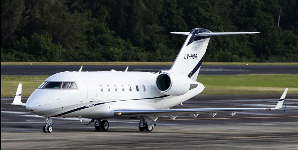 A white business jet with dark blue and grey stripes on the tail and fuselage, registration LV-HQR, is parked on a tarmac with trees in the background.
