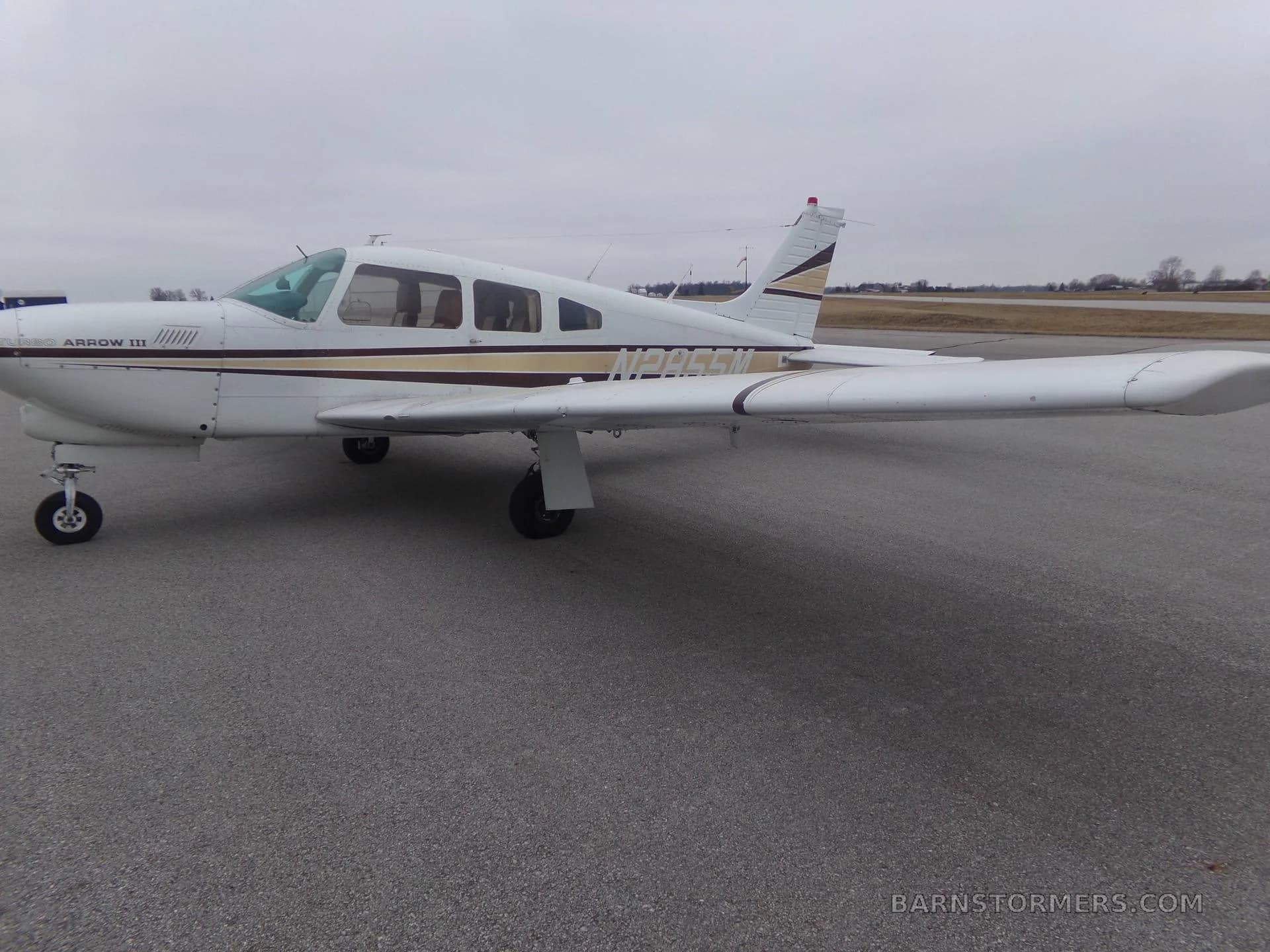 A white Piper Arrow III aircraft with brown and gold stripes, registration N2855M, is parked on an asphalt tarmac, viewed from the right side.