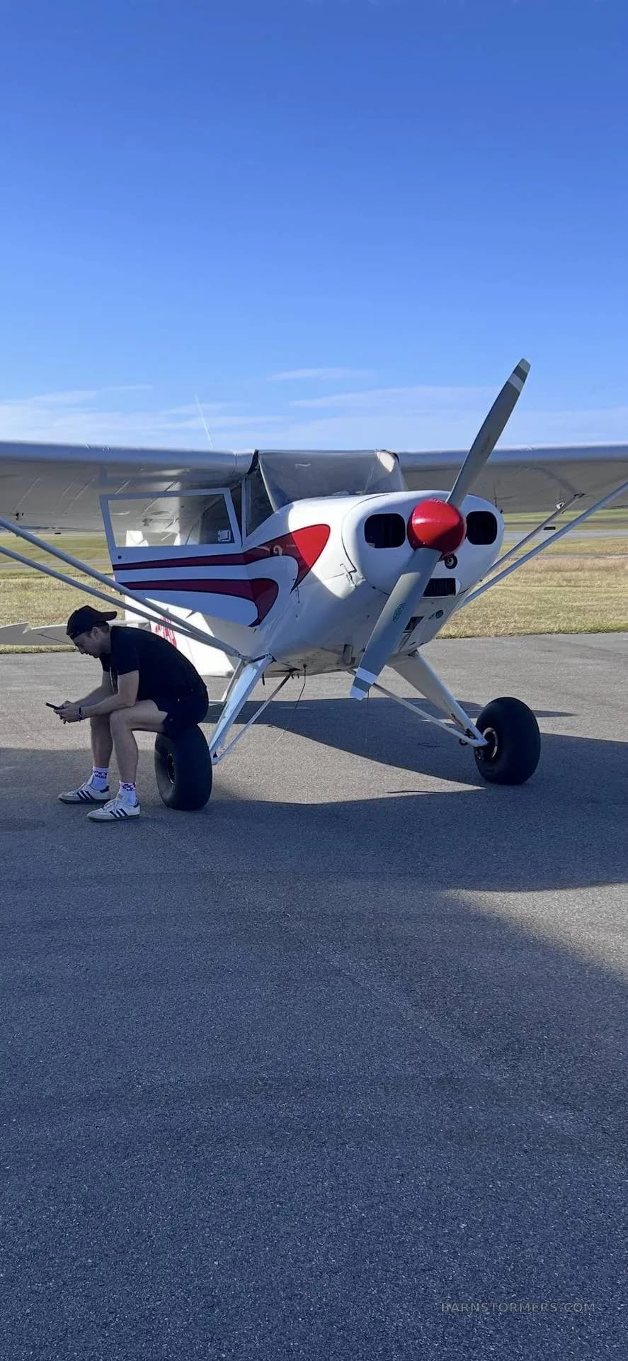 A white single-engine aircraft with red stripes on the tarmac, with a person sitting on its wheel.