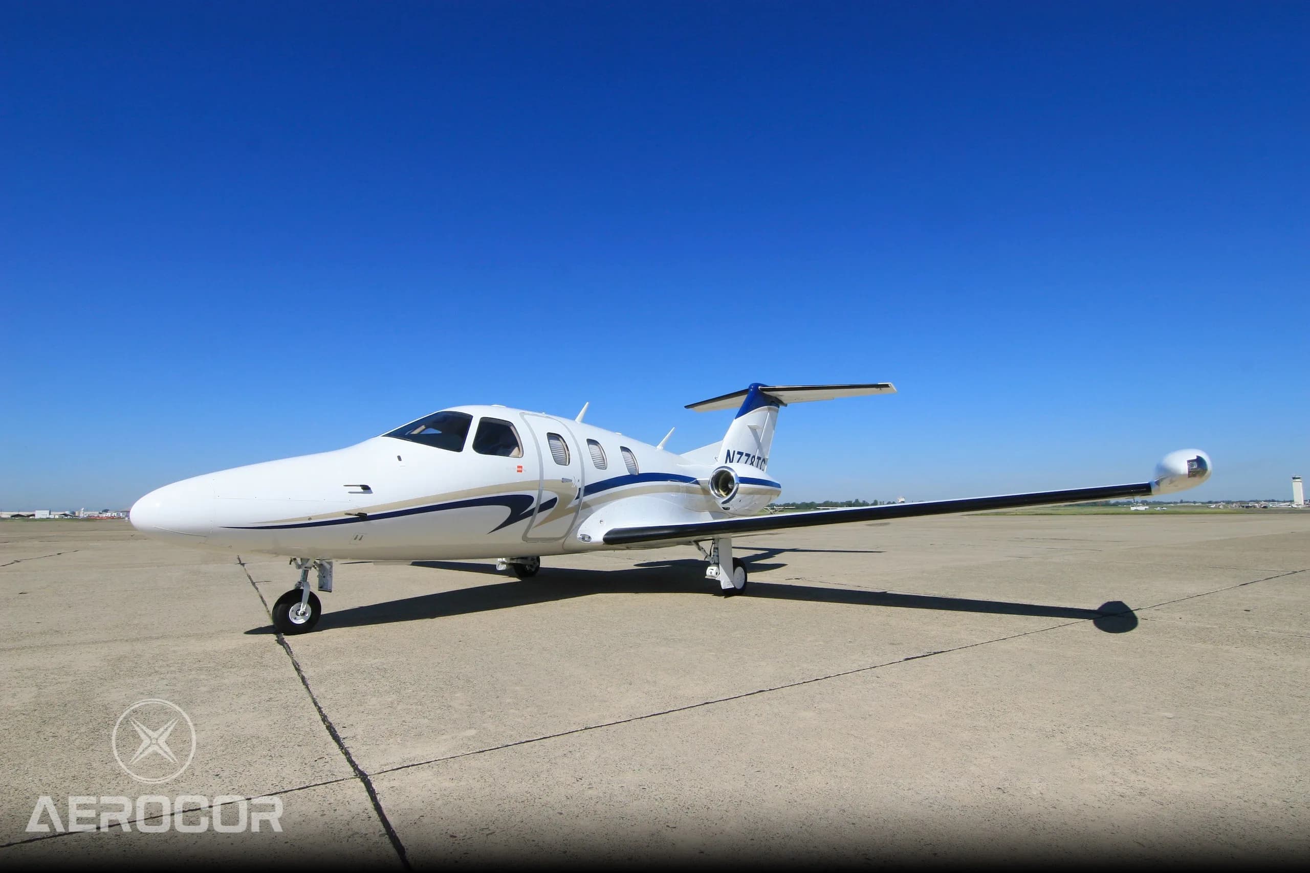 A white Eclipse 500 jet with blue and gold stripes on the tarmac under a clear blue sky.