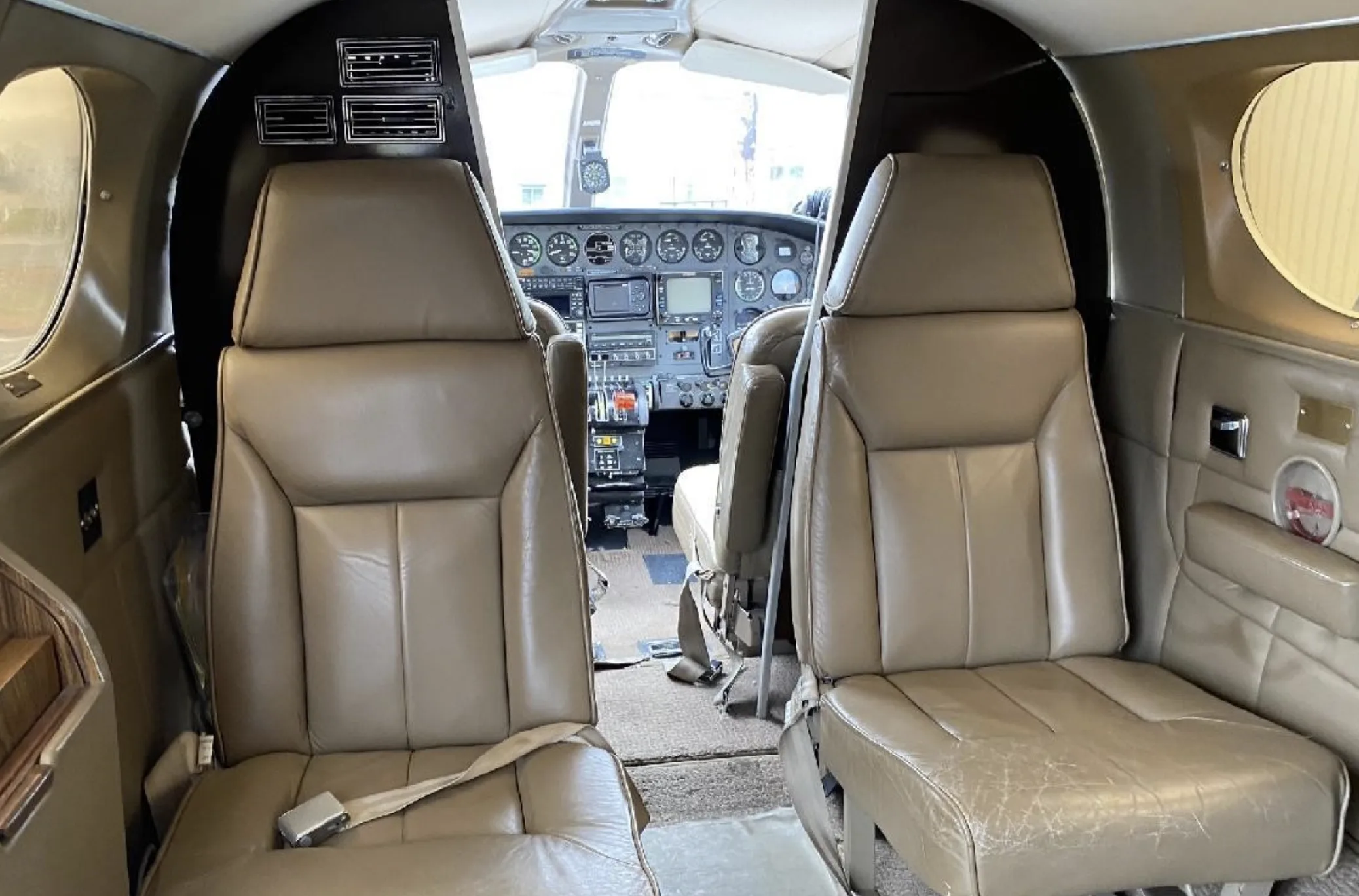 The aircraft's cabin interior features two tan leather seats in the foreground, looking forward towards the cockpit.