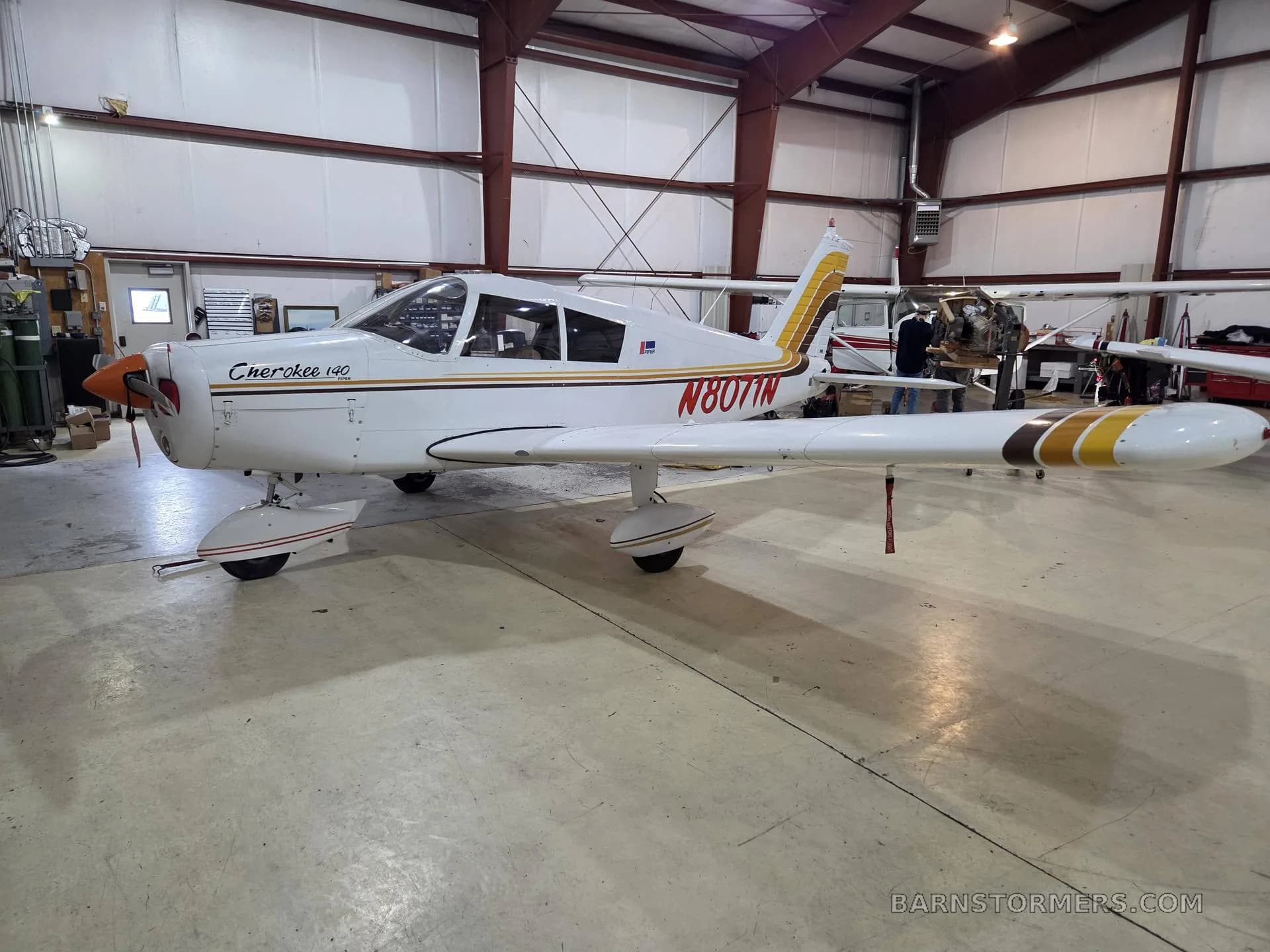 A side view of a white Piper Cherokee 140 with brown and gold stripes, registration N8071N, in a hangar.