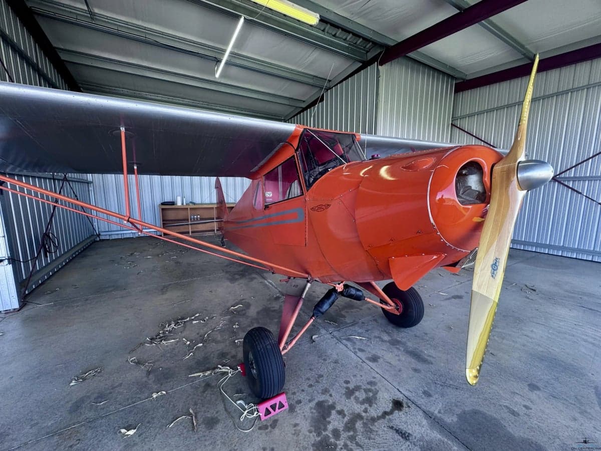 A wide front-right view of the orange vintage aircraft with a wooden propeller inside a hangar.