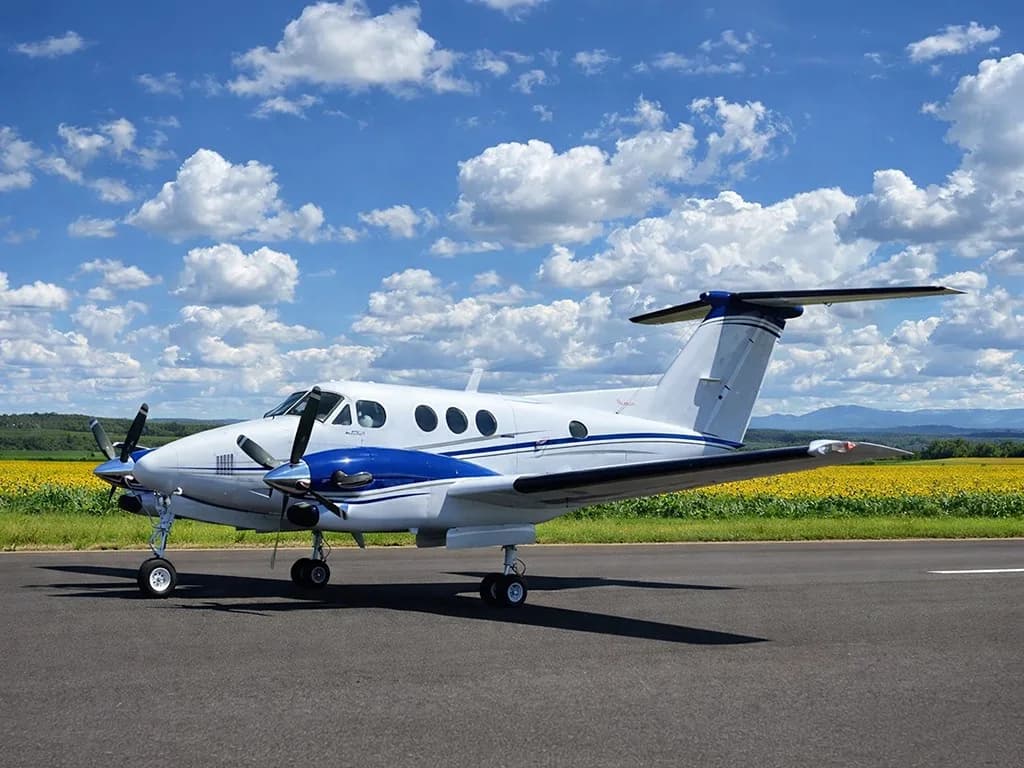 A white and blue twin-engine turboprop aircraft on a runway with a field of yellow flowers and blue sky in the background.