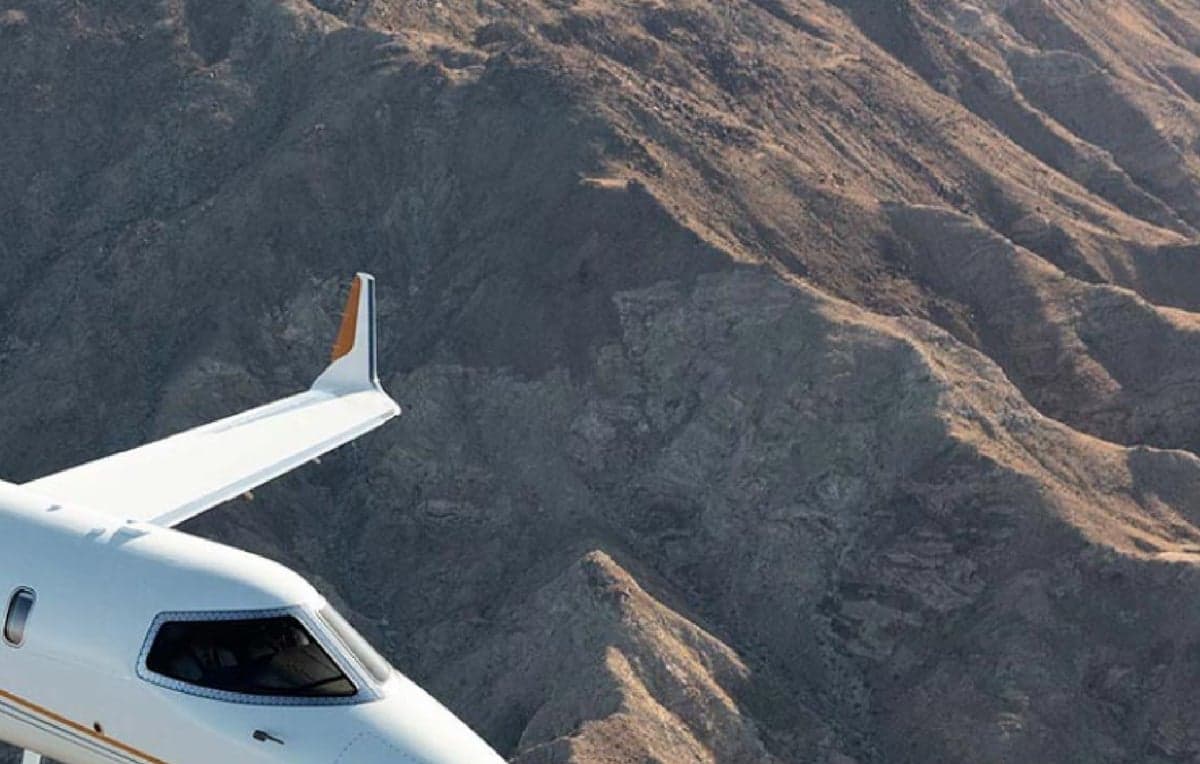 Partial view of an aircraft wing and cockpit flying over a mountainous landscape.