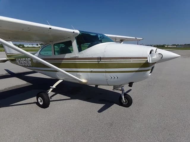 A white Cessna 182 with olive green and brown stripes, registration N2850F, parked on an asphalt tarmac under a clear blue sky.
