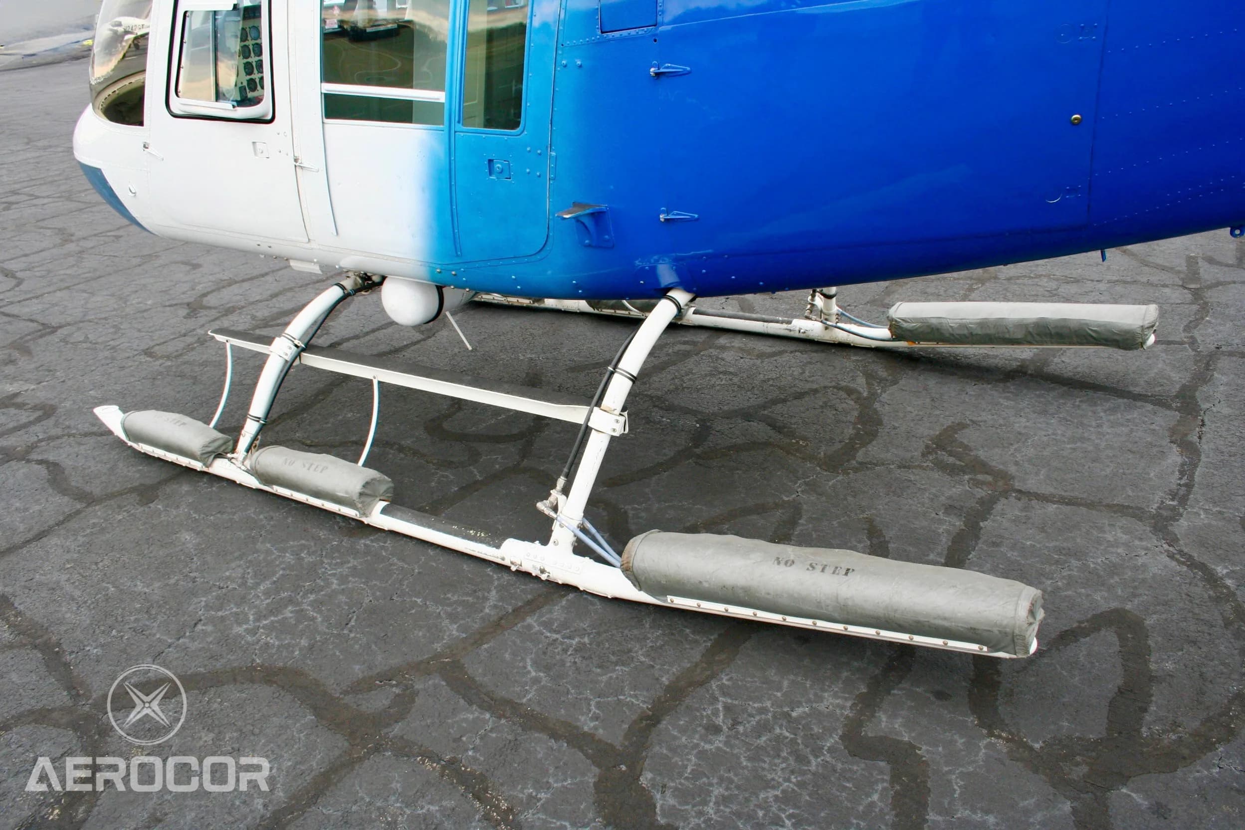 A close-up view of the skid landing gear of the blue and white Bell JetRanger III helicopter, showing "NO STEP" markings on the floats.