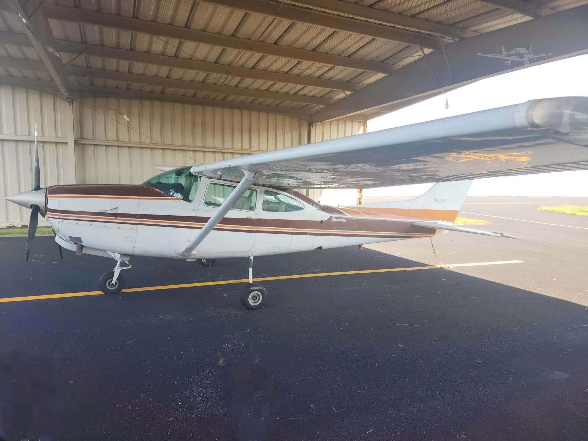 A white Cessna 182 Skylane II with brown and orange stripes is parked under a hangar roof on an asphalt ramp.