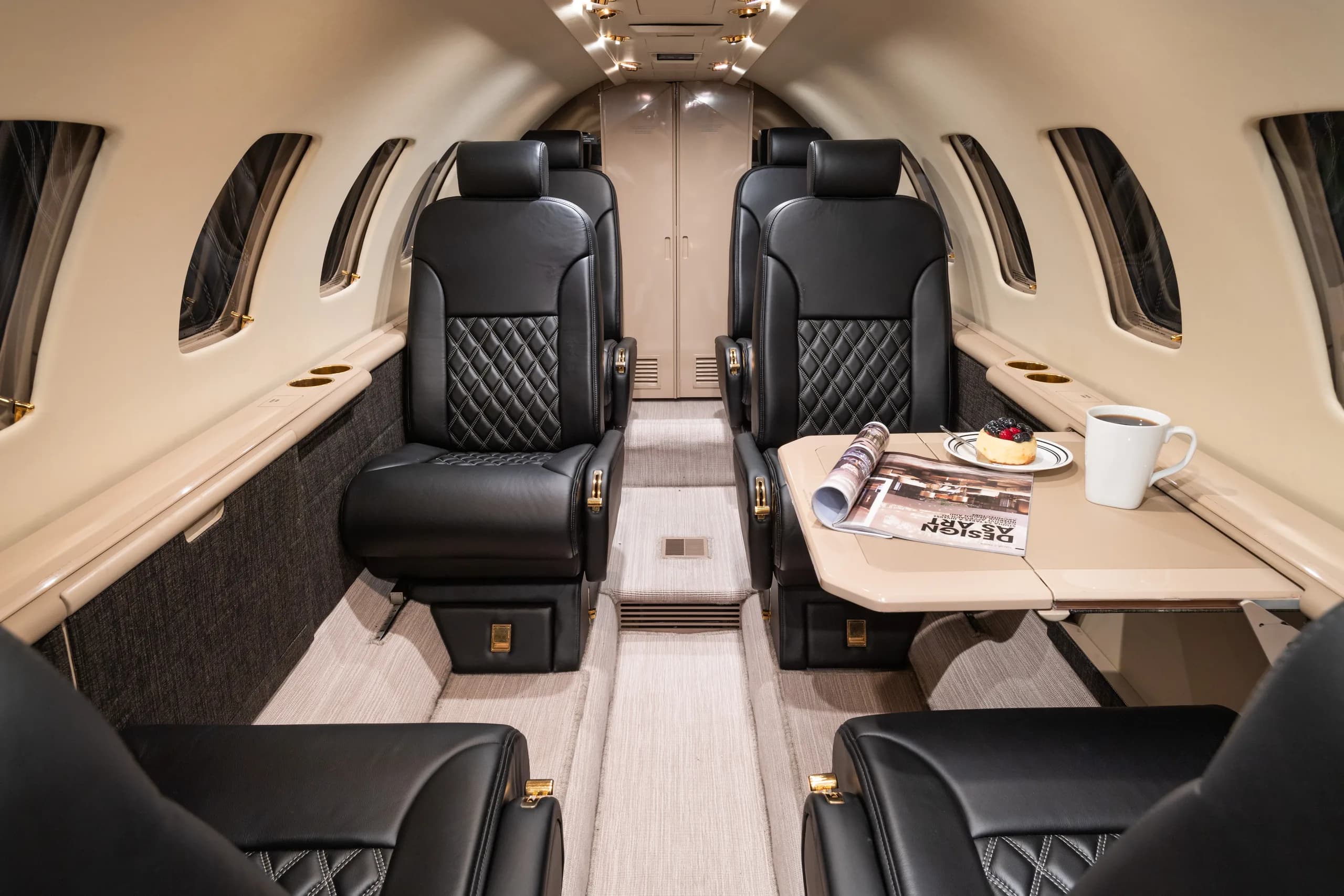 The luxurious interior cabin of a business jet with black quilted leather seats and a light-colored table set with a magazine, coffee, and pastry.