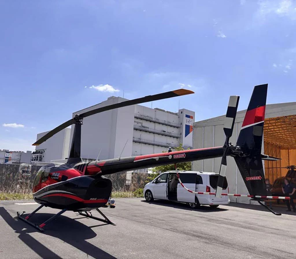 A black and red Robinson R66 Turbine helicopter is parked on an airfield next to a white van and large buildings.