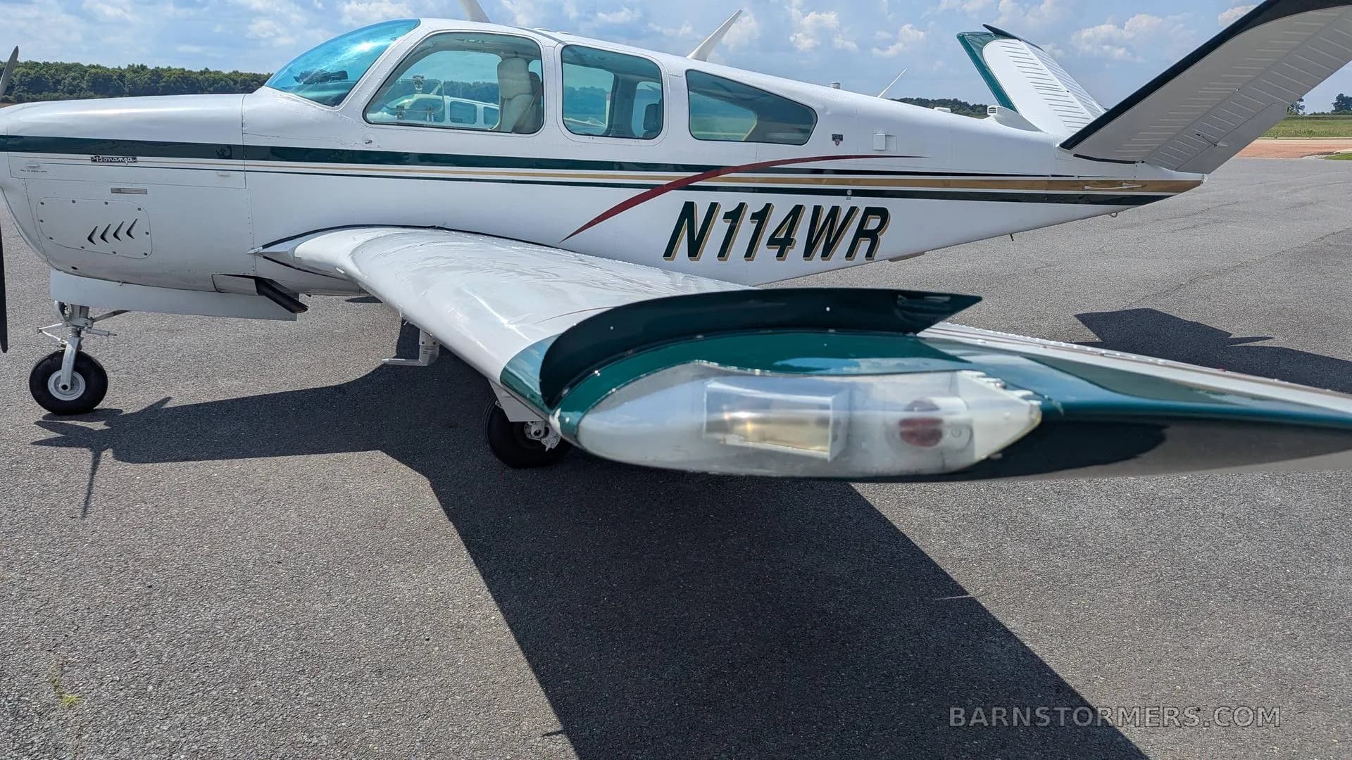 A full side view of the white Beechcraft Bonanza N114WR, with green and gold stripes, parked on an asphalt ramp.