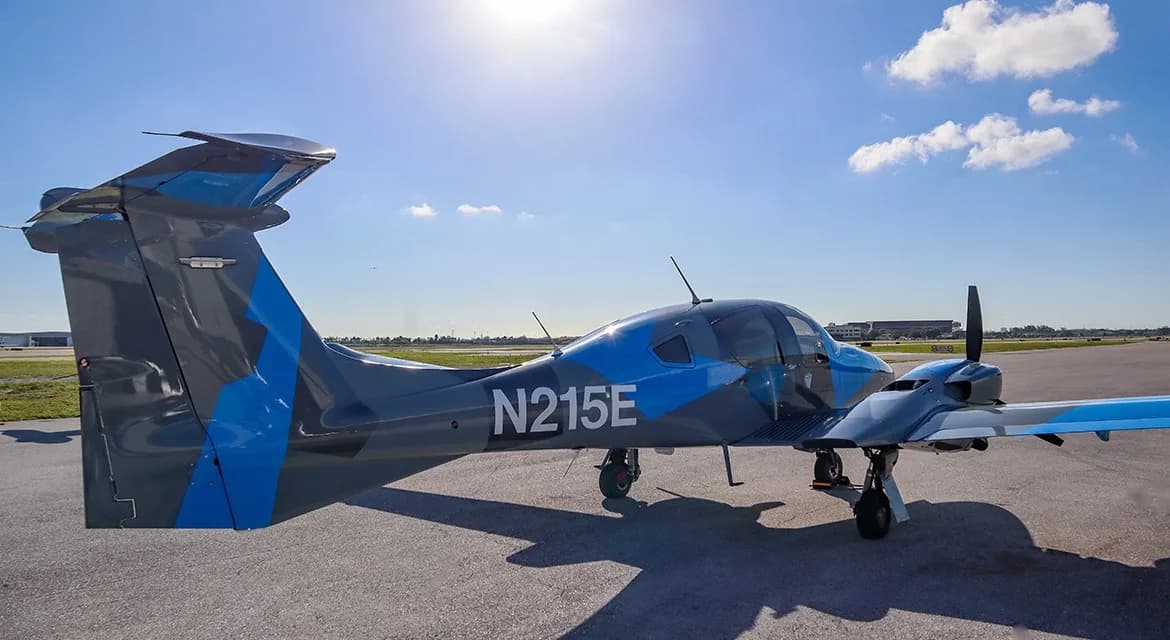 A rear-side view of a gray and blue Diamond DA62 aircraft, registration N215E, parked on an airfield under a sunny sky.