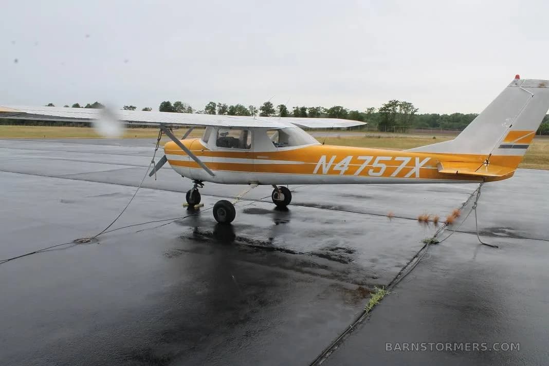 A yellow and white Cessna 150 (N4757X) is parked on a wet tarmac, viewed from the rear-left.