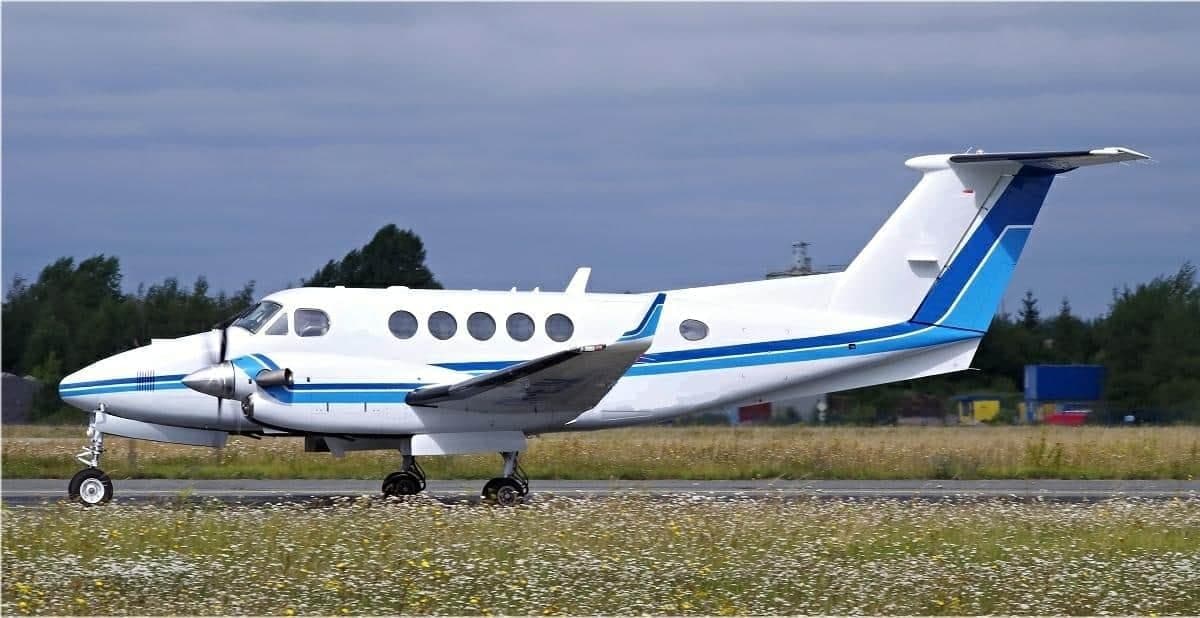 A white and blue Beechcraft King Air turboprop aircraft taxiing on a runway next to a field of wildflowers under a cloudy sky.