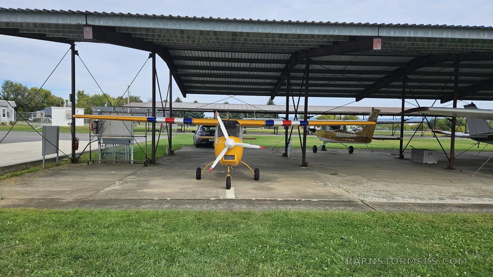 A yellow Merlin ultralight aircraft with a red, white, and blue striped wing is viewed from the front, parked under an open-sided hangar.