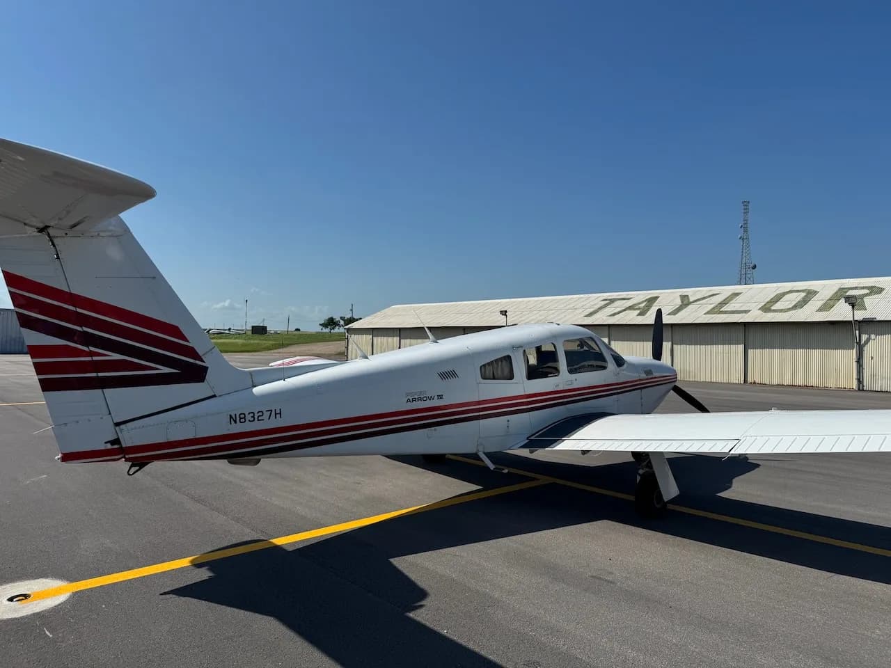 A white Piper Arrow II aircraft with red and brown stripes parked on an asphalt tarmac, viewed from the right side.