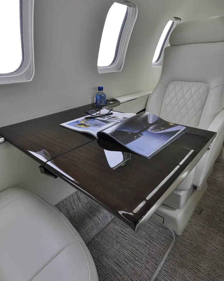 Aircraft interior with a white leather seat, a dark wood table with a magazine, and a bottle of water.