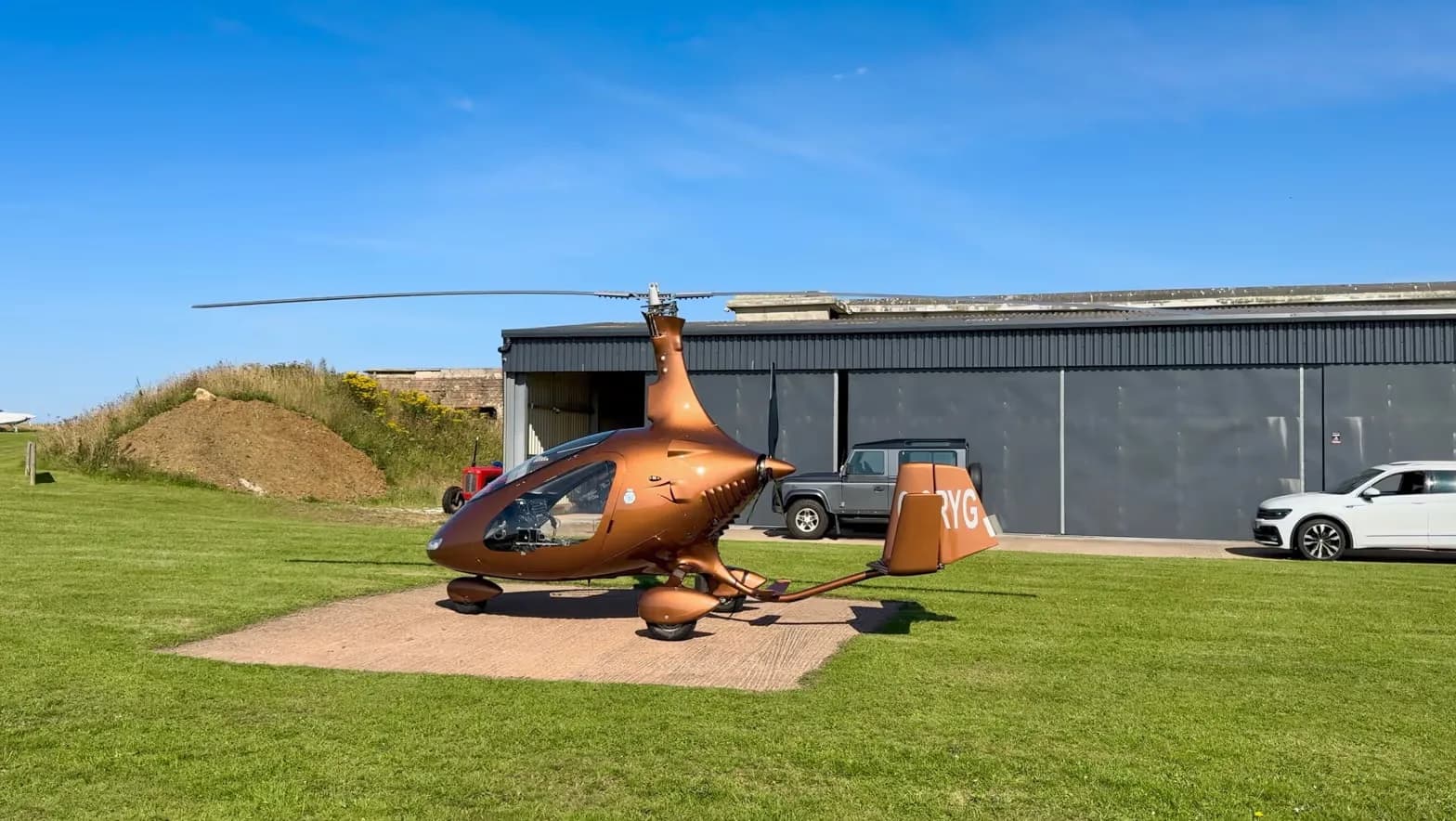 A brown gyrocopter, G-ORYG, parked on a concrete pad in front of a hangar with other vehicles.