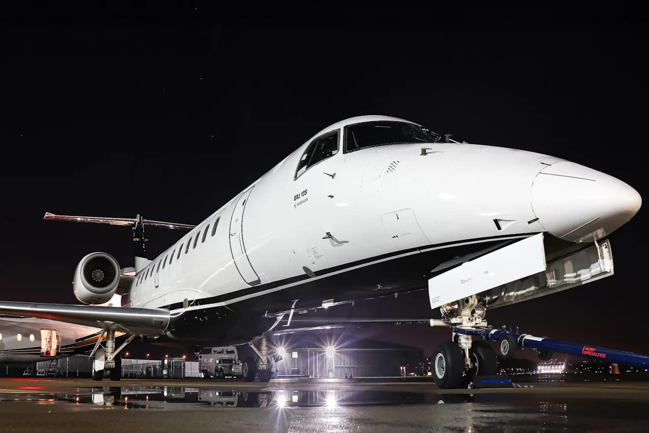 A white and black Embraer ERJ 135 aircraft is parked on a wet tarmac at night, illuminated by ground lights.