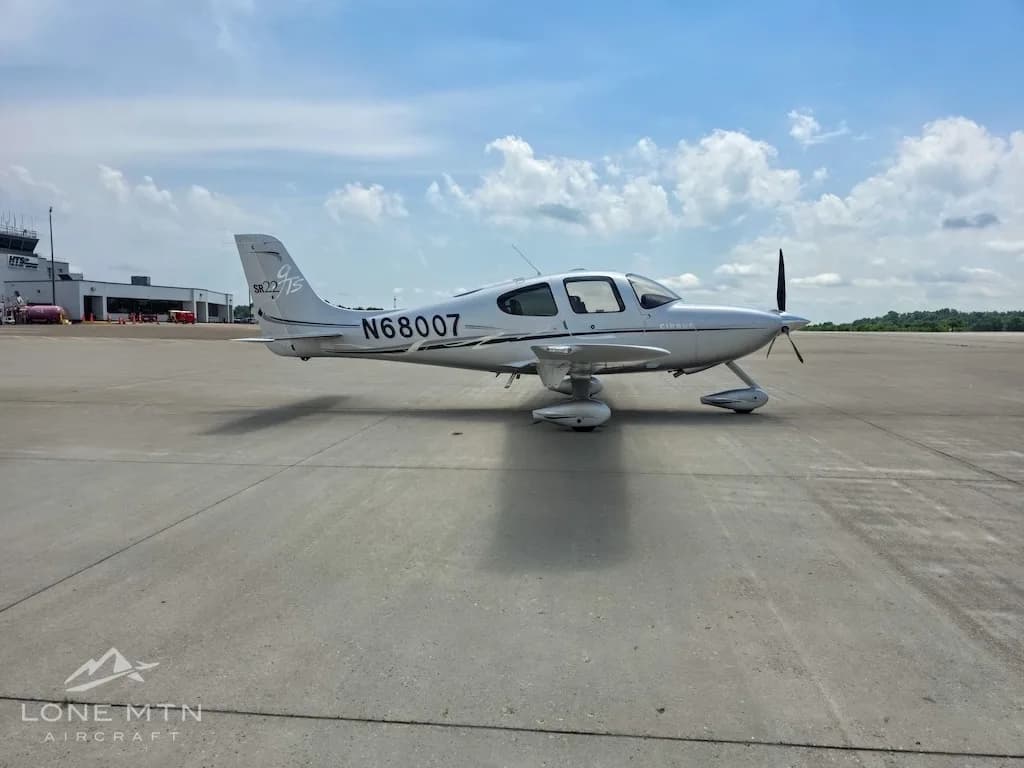 A silver Cirrus SR22 aircraft (N68007) with its doors closed, parked on an airfield with airport buildings in the background.